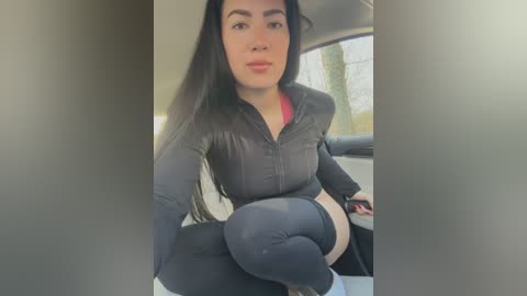 Video of a pregnant woman with long black hair, wearing a black zip-up jacket and black leggings, seated in a car, with a blurred background.