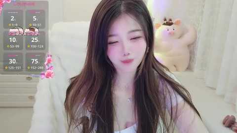 A young Asian woman with long black hair and fair skin, wearing a white top, sits in a softly lit room. A plush pig toy is visible behind her.