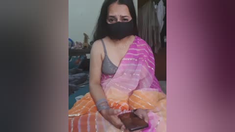 Media: A video of a South Asian woman with long black hair, wearing a gray bra, pink and orange sari, and a black face mask, sitting on a bed in a dimly lit room, holding a smartphone.