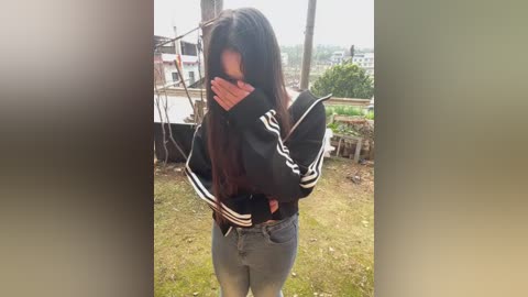 Video of a young woman with long, dark hair, covering her face with her hands, wearing a black hoodie and jeans, standing outdoors near a wooden structure and greenery.