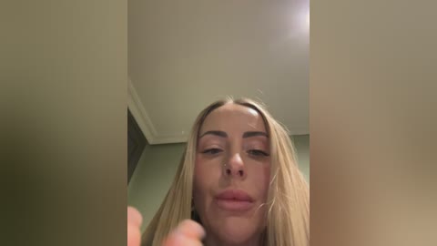 Video of a young woman with long blonde hair, pale skin, and full lips, taken from a low angle, showing her from the neck up in a dimly lit room with beige walls.