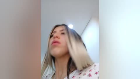 A video of a young woman with light skin and shoulder-length blonde hair, wearing a floral-patterned top, standing indoors against a plain white wall.