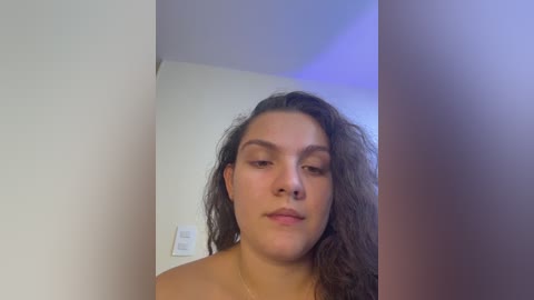 Video of a young woman with medium skin tone and curly brown hair, topless, standing in a dimly lit room with a white wall and a light switch.