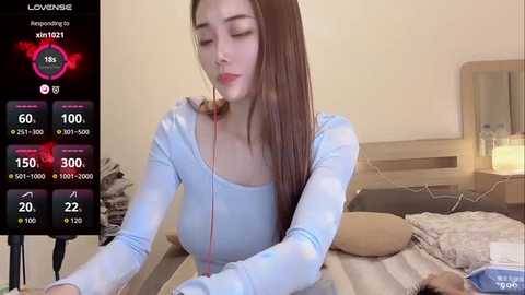 A video of a young woman with long, straight brown hair, wearing a light blue top, sitting on a bed with a screen displaying \"Live Streaming\" and \"Viewer Count\" in the background.