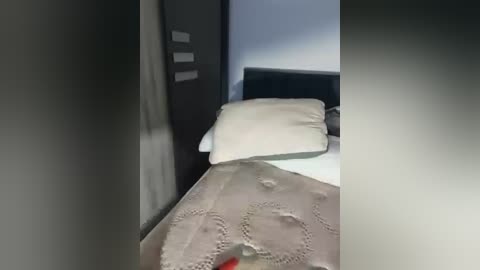 Media: A video of a neatly made bed with a beige quilted bedspread, white pillows, and a black headboard. The room is dimly lit, with a dark wall on the left and a light-colored wall on the right.