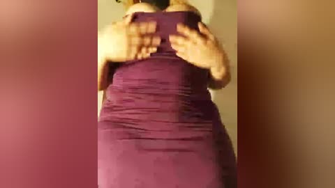Video of a person from behind, wearing a snug, deep purple dress, with their hands placed on their shoulders, creating shadow patterns on the wall. Background is blurred.