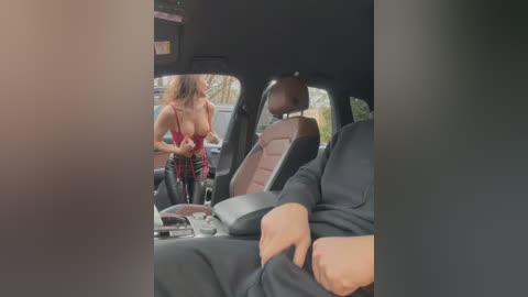 Video of a woman in a red corset and plaid skirt, standing in a car next to a man in a black suit, both in a modern, well-lit setting.