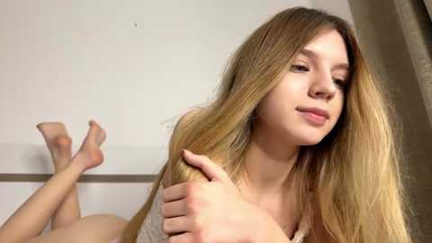 A young, light-skinned woman with long, straight blonde hair, lying on a bed, holding her hair, legs crossed, wearing a white tank top.