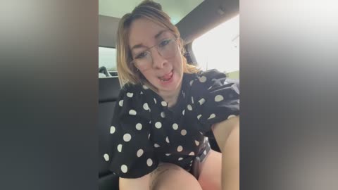 Video of a woman with light skin, shoulder-length blonde hair, wearing glasses, a black polka-dot dress, and a playful expression, sitting in a car, with a blurred background.