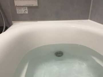 Video of a modern, white bathtub with a clear, calm water surface, surrounded by light grey tiled walls, featuring a small, round drain in the center. The image focuses on the bathtub's interior, with a subtle, clean, and minimalist aesthetic.