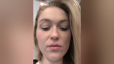 Video of a young Caucasian woman with light skin and straight, blonde hair, wearing a nose ring. She has a neutral expression with slightly parted lips. The background is blurred and beige.