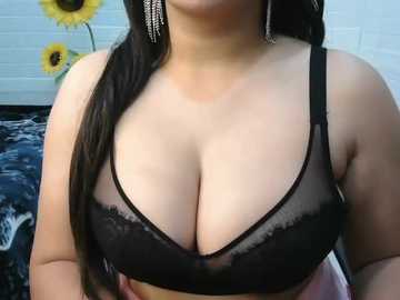 Video of a light-skinned woman with long dark hair, wearing a sheer black lace bra revealing ample cleavage. Background features a tiled wall with sunflowers and a black and white abstract painting.