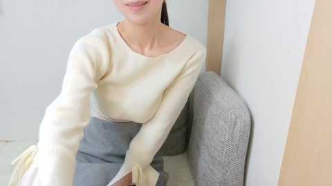 Video of a light-skinned woman with long brown hair, smiling, wearing a cream-colored long-sleeve sweater and high-waisted gray skirt, seated on a gray cushioned chair in a minimalist, neutral-toned room.