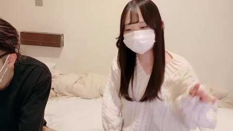 Video of a young East Asian woman with long black hair, wearing a white mask and a cream-colored sweater, gesturing with her right hand in a cozy, beige-toned bedroom.