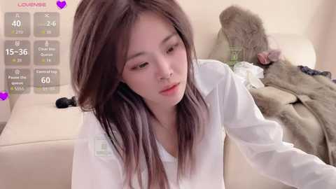 Video of a young Asian woman with long, straight black hair, wearing a white long-sleeve shirt, sitting on a beige couch. Background includes a brown fur coat, a white dress, and a purple heart overlay.