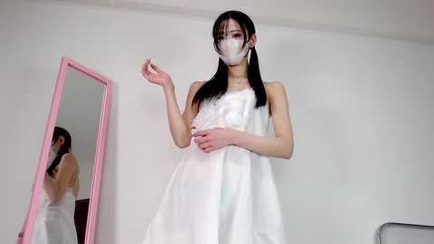 Video of an Asian woman with straight black hair, wearing a white dress, face mask, and holding a phone, in front of a pink-framed mirror.