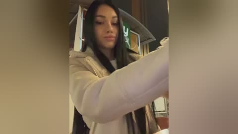 A video of a young woman with long dark hair, wearing a beige coat and scarf, adjusting a poster in an office.