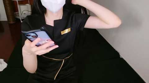 Video of a woman with long black hair, wearing a black medical uniform with gold trim, white mask, and holding a smartphone, standing in a dimly lit room with a green mat.