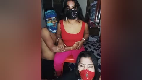 Video of a South Asian woman with medium skin tone and black hair, wearing a red mask and red pants, sitting on a bed, with a man's head wrapped in a colorful turban beside her.