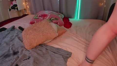 Video of a messy bedroom with a woman in the background, wearing a gray shirt and a bracelet, sitting on a bed with beige sheets, pink floral pillows, and a fuzzy blanket.