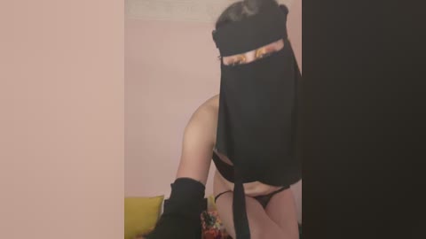 Video of a person in black lingerie, black headband, and black knee-high socks, partially obscured by a partition, standing in a room with pink walls and a green couch.