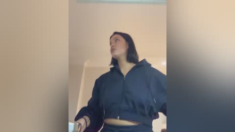 A video of a woman with shoulder-length black hair, wearing a navy blue crop top and high-waisted pants, standing in a beige hallway, looking upwards.