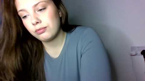 Video of a young woman with long brown hair, wearing a light blue shirt, leaning forward with a neutral expression against a plain white wall.