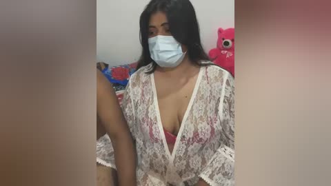 Video of a dark-skinned woman with long black hair, wearing a white lace robe, a pink bra, and a blue mask, sitting in a room with a pink teddy bear and a blue blanket.