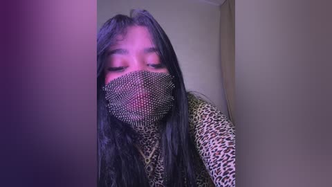 Video of a woman with long, dark hair, wearing a leopard print mask and a leopard print top, against a beige wall, bathed in purple lighting.