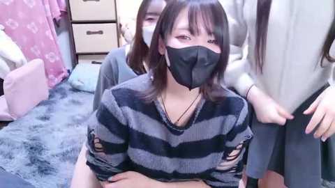 Video of three Asian women in a bedroom, one with a face mask, striped shirt, and torn sleeves, another with long black hair, and a third partially visible.