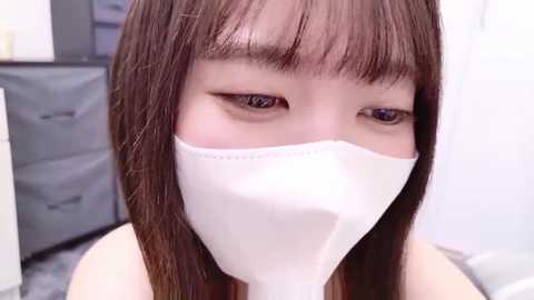 Video of an Asian woman with straight brown hair, wearing a white surgical mask, looking down. Background shows a blurred, grey metal cabinet and a white wall.