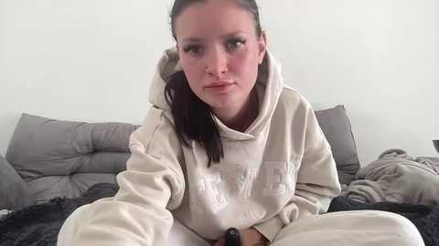 Video of a young Caucasian woman with fair skin, long brown hair, and green eyes, wearing a beige hoodie, sitting on a bed with gray pillows and a black blanket.