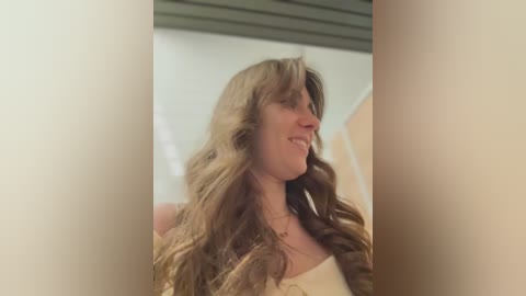 Video of a smiling woman with long, wavy, light brown hair, wearing a white tank top, taken indoors with blurred background featuring a white ceiling and wall.