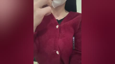 Media: Video of a person wearing a red cardigan, white face mask, and light-colored shirt, with their left hand near their face. The background is blurred in a reddish hue, obscuring details.