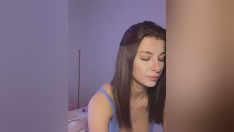 Video of a young woman with long brown hair, wearing a blue spaghetti-strap top, leaning against a cylindrical pillar in a dimly lit room with soft purple lighting.