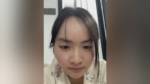 Video of an Asian woman with medium-length black hair, wearing a cream blouse with floral patterns, looking down with a neutral expression. Background features a white wall and a black-framed window.