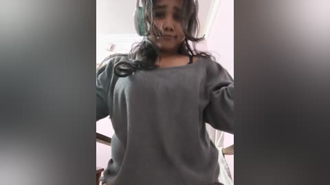 A video of a young, dark-skinned girl with curly hair, wearing a grey sweatshirt and headphones, standing indoors with blurred background.