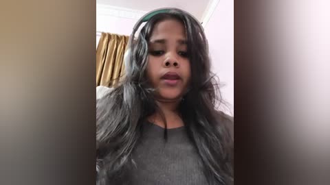 A video of a young Black girl with long, wavy black hair, wearing a gray shirt, standing in a room with light pink walls and a brown curtain in the background.