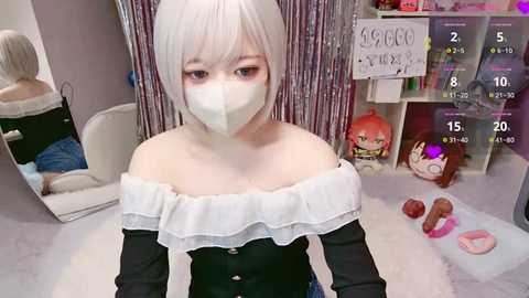 Video of a young woman with white hair in a cosplay outfit, wearing a mask and off-shoulder black top, standing in a cluttered room with a mirror and toy shelves.