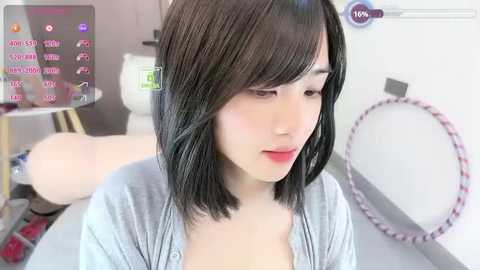 Video of an Asian woman with shoulder-length black hair, fair skin, and a slender build, wearing a light gray cardigan. Background includes a pink hula hoop, white wall, and a digital sign displaying \"Vtuber\" text.