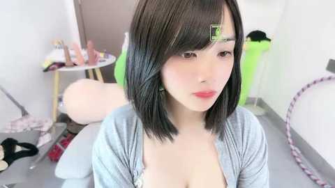 Video of a young Asian woman with straight black hair, light skin, wearing a light gray cardigan, sitting in a cluttered room with a green chair, pink bicycle, and makeup items scattered on the floor.