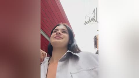 Media: Video of a young woman with fair skin and straight, dark hair, wearing a light gray denim jacket, standing in a modern, white-walled room with red brick wall and industrial-style lighting.