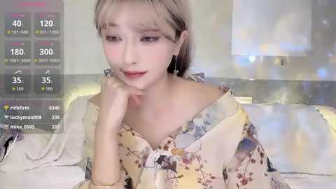 Video of a young Asian woman with light skin and blonde hair, wearing a floral blouse, sitting on a bed. Background shows a blurred bedroom with white walls and a digital overlay with social media statistics.