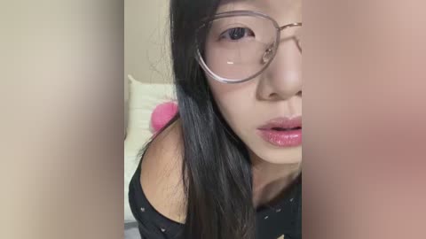 A close-up video of an Asian woman with long black hair, wearing glasses, pink lipstick, and a black off-shoulder top, partially blurred, against a beige wall with a white pillow.