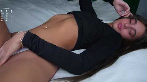 Video of a young, light-skinned woman with long brown hair, lying on a bed in a black long-sleeve crop top and white panties, her eyes closed and hand on her stomach.