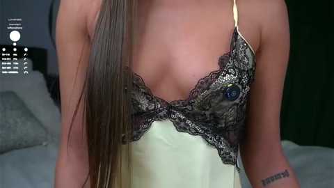 Video of a woman in a pale yellow satin camisole with black lace trim, revealing her medium-sized breasts, against a blurred dark green background.