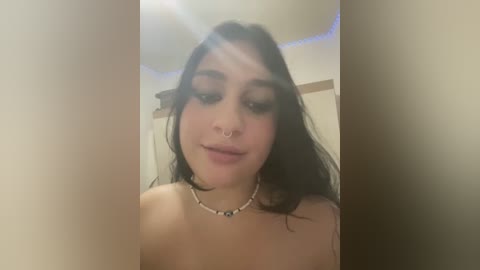 Media: Video of a topless woman with long dark hair, multiple piercings, and a silver necklace, indoors with a blurred background.