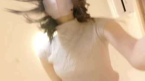 A blurry video of a woman with wavy brown hair, wearing a white ribbed sweater, captured mid-motion against a beige wall.