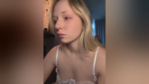 Video of a young, fair-skinned blonde woman with straight hair, wearing a white lace bra, looking contemplative indoors.