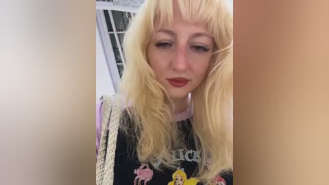 Video of a young woman with long, straight, platinum blonde hair and bangs, wearing a black graphic t-shirt featuring a yellow character. She has fair skin, red lipstick, and is indoors near a window with a white frame.
